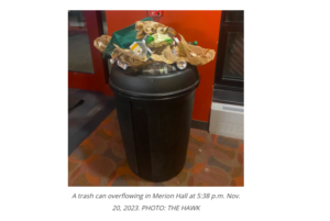 A black trash can sits on a reddish brown carpet in Merion Hall. There is a a red wall behind it, a window to the right of it and a door to the left of it. The trashcan is overflowing with various pieces of garbage piled up over the top of the can. The Alt Text under the photo reads, "A trash can overflowing in Merion Hall at 5:38 p.m. Nov. 20, 2023. PHOTO: THE HAWK."