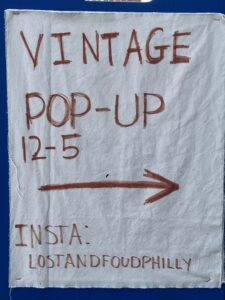 Photo of a vintage pop up store entrance sign. The sign has a white cloth background with red marker writing. The sign reads: "Vintage Pop-Up, 12-5, Insta:LostandFoundVintage"
